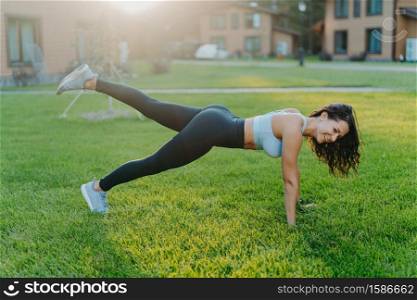 Happy sporty woman stands in plank pose, raises legs and does physical exercises to keep fit, dressed in top and leggings, goes in for sport regularly, enjoys nice summer day, poses on green grass