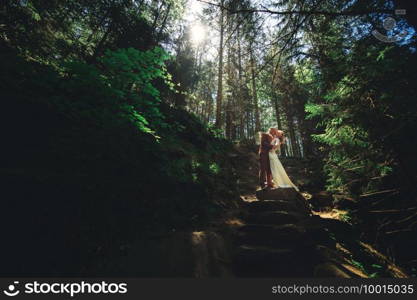 Happy stylish couple newlyweds in the green forest on summer day. bride in long white dress and groom in red suit are hugging. wedding day.. Happy stylish couple newlyweds in the green forest on summer day. bride in long white dress and groom in red suit are hugging. wedding day
