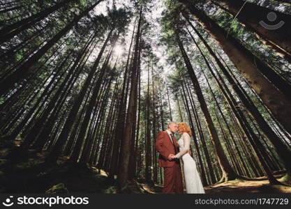 Happy stylish couple newlyweds in the green forest on summer day. bride in long white dress and groom in red suit are hugging. wedding day.. Happy stylish couple newlyweds in the green forest on summer day. bride in long white dress and groom in red suit are hugging. wedding day
