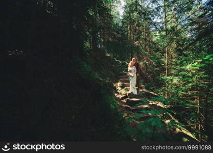 Happy stylish couple newlyweds in the green forest on summer day. bride in long white dress and groom in red suit are hugging. wedding day. Happy stylish couple newlyweds in the green forest on summer day. bride in long white dress and groom in red suit are hugging. wedding day.