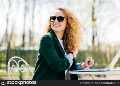 Happy stylish woman with curly light hair wearing sunglasses working with laptop outside in park typing necessary documents turning back noticing someone at the street. Cheerful woman having rest