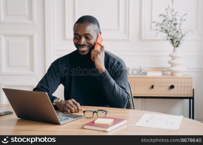 Happy successful african american businessman having pleasant telephone conversation while working on laptop at home office, smiling dark skinned male employee discussing project with partner by phone. Successful afro american businessman having pleasant telephone conversation while working on laptop