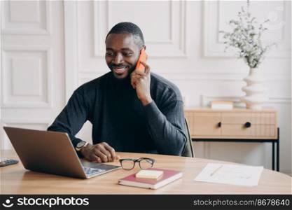 Happy successful african american businessman having pleasant telephone conversation while working on laptop at home office, smiling dark skinned male employee discussing project with partner by phone. Successful afro american businessman having pleasant telephone conversation while working on laptop
