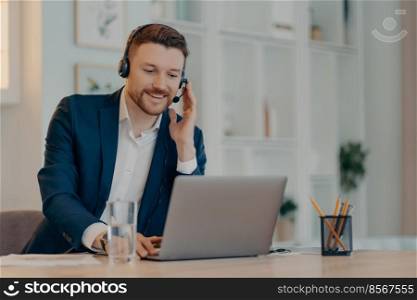 Happy successful businessman using laptop and typing on keyboard while sitting at the table and doing video call with headset, taking part in web conference. Online business concept. Young man in suit talking on webcam conference on laptop at home