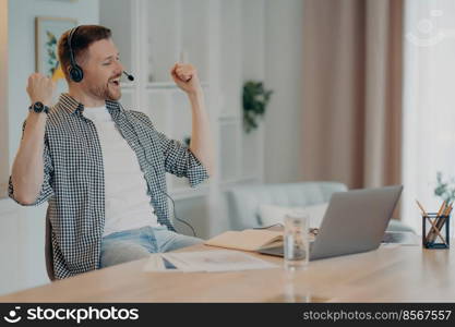 Happy successful man clenches fists celebrates triumph works online wears headset poses at desktop with laptop computer dressed casually. Overjoyed male student listens educational online lecture