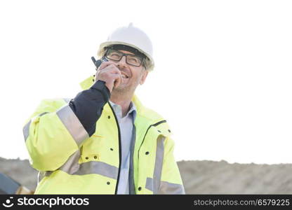 Happy supervisor using walkie-talkie at construction site against clear sky