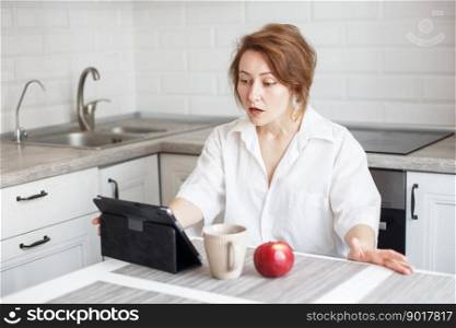 Happy surprised woman with cup of coffee or tea using laptop in quarantine lockdown in the kitchen at home in the white shirt. girl with a tablet. Happy surprised woman with cup of coffee or tea using laptop in the kitchen in white shirt. girl with a tablet