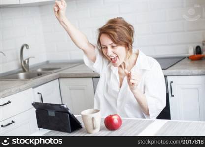 Happy surprised woman with cup of coffee or tea using laptop in quarantine lockdown in the kitchen at home in the white shirt. girl with a tablet. Happy surprised woman with cup of coffee or tea using laptop in the kitchen. girl with a tablet raised her hands up