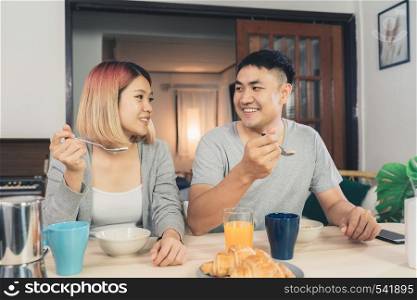 Happy sweet Asian couple having breakfast, cereal in milk, bread and drinking orange juice after wake up in the morning. Husband and his wife eating food together.