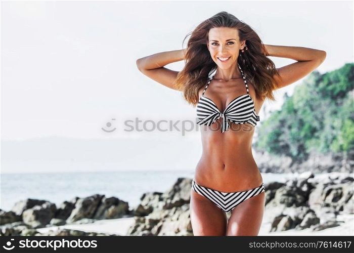 Happy tanned girl in bikini at seaside, blue sea and stony beach on background. Girl in bikini at seaside