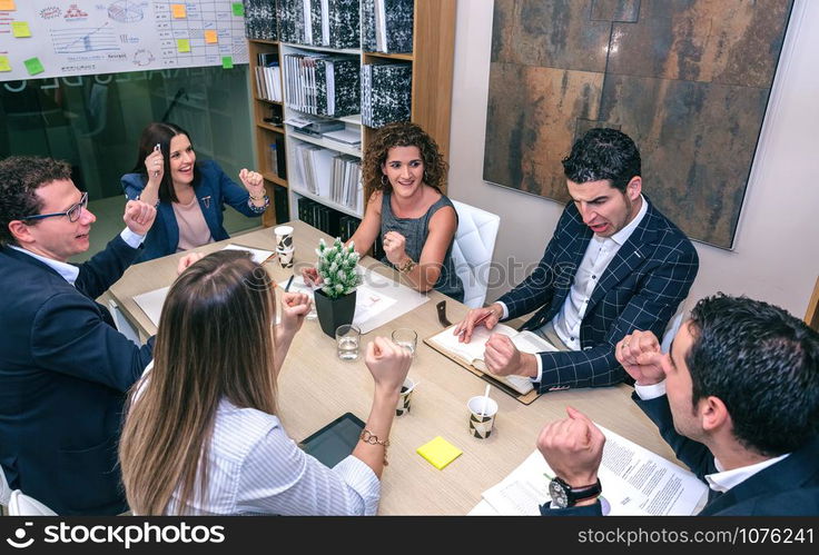 Happy teamwork celebrating business success with hands up sitting around of table in company headquarters. Teamwork celebrating business success in company headquarters