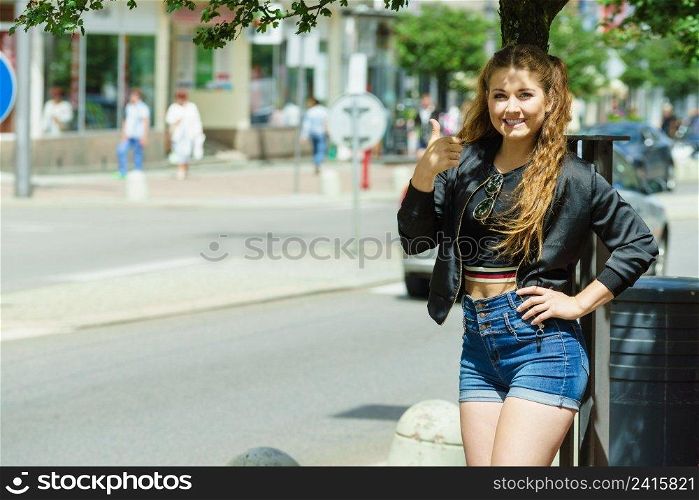 Happy teen woman enjoying her leisure time in city. Female having long brown hair in ponytails wearing black leather jacket and denim shorts. Happy beautiful young teenager woman