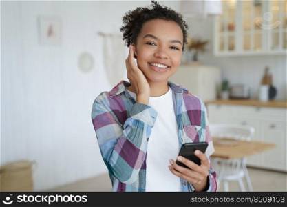 Happy teenage afro girl enjoying music in wireless earphones on wifi. Music podcast listening in airpods on mobile phone. Rest and recreation at home. Attractive young woman is having fun.. Happy teenage girl enjoying music podcast listening in wireless earphones. Recreation at home.