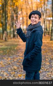 Happy teenage boy in the autumn sunny park. Happy smiling teenage boy in a beret and jacket waves his hand in greeting in the autumn sunny park