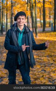Happy teenage boy in the autumn sunny park. Happy smiling teenage boy in a beret and jacket showing gesture to the right in the autumn sunny park