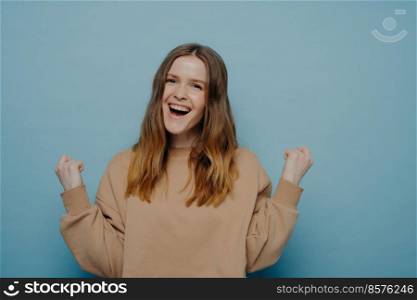 Happy teenage girl feeling satisfied after accomplishing her goal or winning important competition, laughing and celebrating success with clenched fists, standing alone next to light wall. Teenage girl celebrating victory or success isolated on blue background