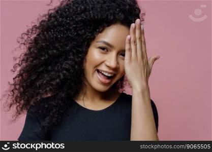Happy teenage girl has fun and hides face with palm, has joyful expression, curly bushy hair, dressed in casual black clothes, isolated on pink background. Shy glad young woman gets compliment