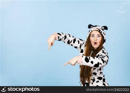 Happy teenage girl in funny nightclothes, pajamas cartoon style pointing down with positive surprised face expression, studio shot on blue. Advertisement concept. Woman wearing pajamas cartoon pointing down