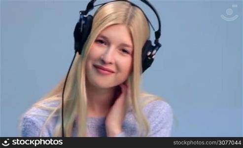 Happy teenage girl in headphones listening to music over white background. Smiling girl in black headphones gently touching her neck, playing with her blonde long hair and smiling at the camera while listening to music track.