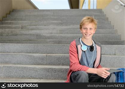 Happy teenage student boy with tablet sitting on steps outdoors