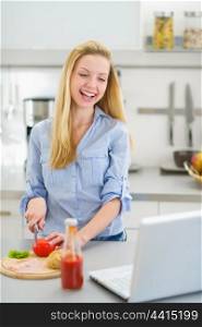 Happy teenager girl making sandwich in kitchen and looking in laptop