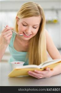 Happy teenager girl reading book and eating yogurt in kitchen