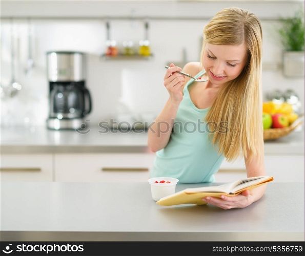 Happy teenager girl reading book and eating yogurt in kitchen
