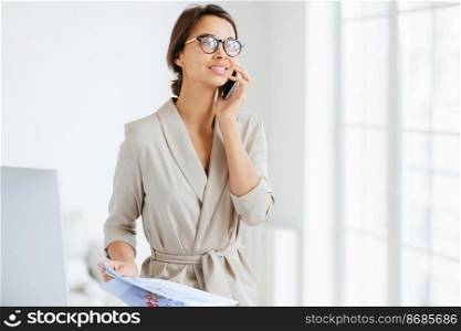 Happy thoughtful successful female employee holds modern cellphone near ear, has telephone conversation, looks aside, dressed elegantly, reviews monthly report, studies graphics, stands in office