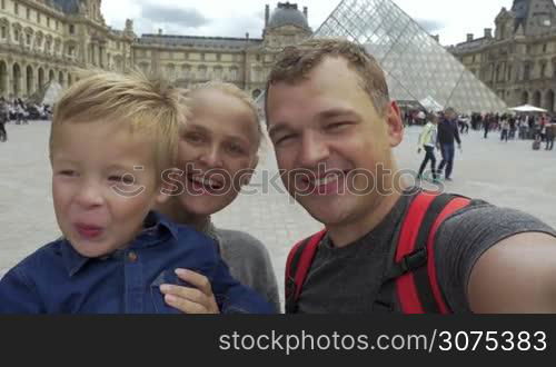 Happy tourists family making selfie video near the The Louvre museum in Paris. Parents smiling and son waving hand to say hello