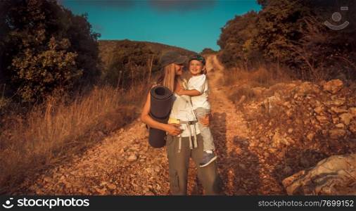 Happy traveler family, active young mother with cute little son on hands walking along mountainous road, enjoying summer c&ing in the mountains