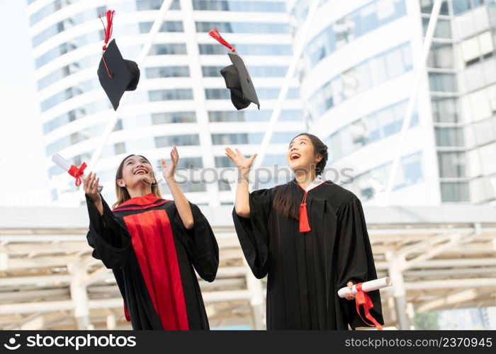 Happy two Asian young beautiful graduate female students with master and bachelor degree throwing cap to the sky after graduation while holding diploma in hand. Blur background of University building