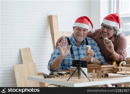 Happy two senior carpenter men Caucasian and asian with beard wearing santa hat selling and showing handmade wooden toys to customers via live on mobile phone at workshop on Christmas holiday