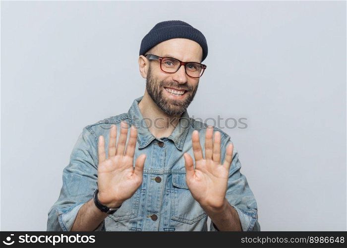 Happy unshaven young male with attractive look shows palms as denies something or demonstrates his refusal, has happy expression, wears stylish black hat, denim jacket and glasses, poses indoor