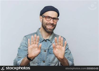 Happy unshaven young male with attractive look shows palms as denies something or demonstrates his refusal, has happy expression, wears stylish black hat, denim jacket and glasses, poses indoor
