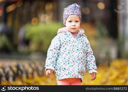 Happy urban little girl walking in city autumn park. Cute baby girl among the golden autumn maple leaves