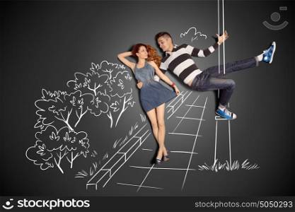 Happy valentines love story concept of a romantic couple against chalk drawings background. Male pole dancing on a lamppost while walking with girlfriend.