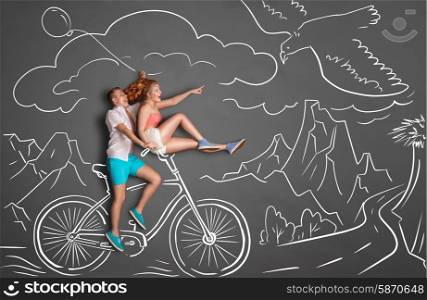 Happy valentines love story concept of a romantic couple on chalk drawings background of mountains. Male riding his girlfriend on a bicycle handle bar, female pointing at a flying eagle.