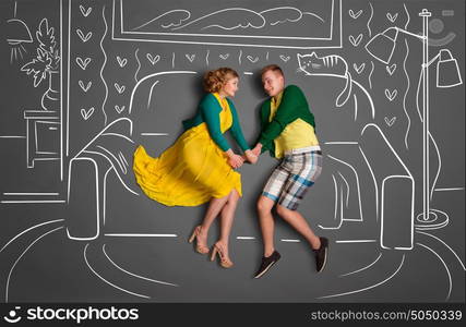 Happy valentines love story concept of a romantic couple sitting on a sofa and holding hands against chalk drawings background of a living room.