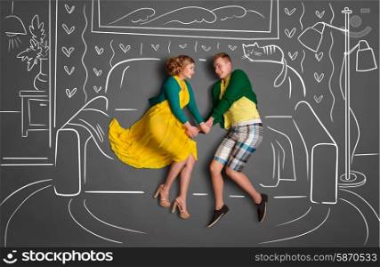 Happy valentines love story concept of a romantic couple sitting on a sofa and holding hands against chalk drawings background of a living room.