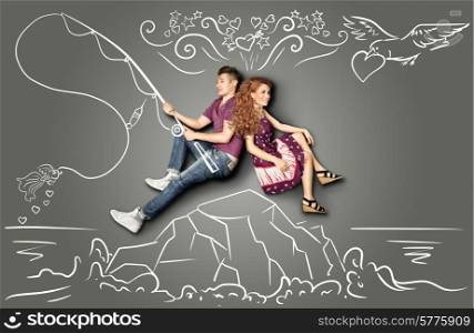 Happy valentines love story concept of a romantic couple sitting on an island and fishing a goldfish on a hook against chalk drawings background.