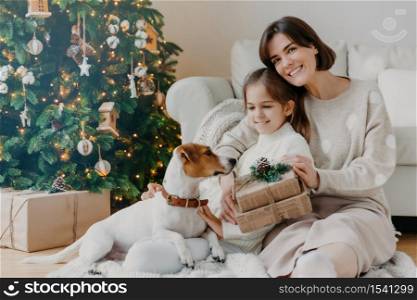 Happy winter holidays! Positive brunette woman embraces little girl pose with gifts on floor in room, jack russell terrier dog near, have fun near Christmas tree. Mom and daughter unpack boxes