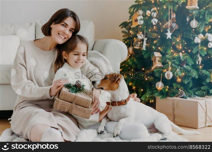 Happy winter holidays  Positive brunette woman embraces little girl pose with gifts on floor in room, jack russell terrier dog near, have fun near Christmas tree. Mom and daughter unpack boxes