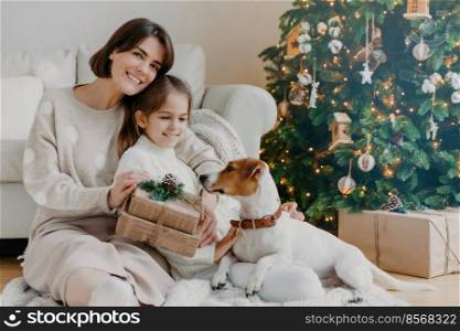 Happy winter holidays  Positive brunette woman embraces little girl pose with gifts on floor in room, jack russell terrier dog near, have fun near Christmas tree. Mom and daughter unpack boxes