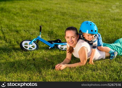 Happy woman and child in helmet having fun outdoor on meadow. Blue balance bike (run bike) laying on grass. Family lifestyle scene of mother and son resting together on green grass in the park.