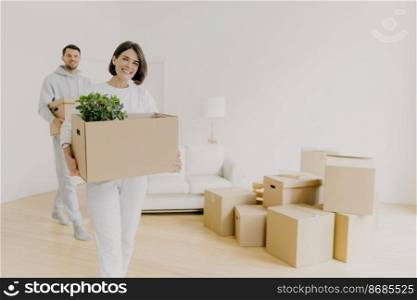 Happy woman and her husband carry boxes with personal belongings, being busy during relocation in other place for living, enter new home, move together in big house, unpack household things.