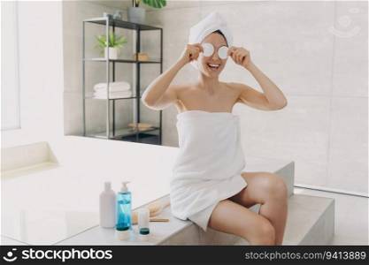 Happy woman applies face toner, has fun with cotton discs. Gorgeous European girl, towel-wrapped post-bath. Lady relaxes on bathtub, showers at home.