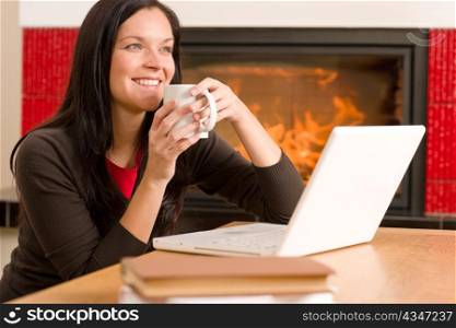Happy woman at fireplace with laptop enjoying winter hot drink