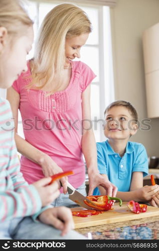 Happy woman chopping red bell pepper while standing with children in kitchen