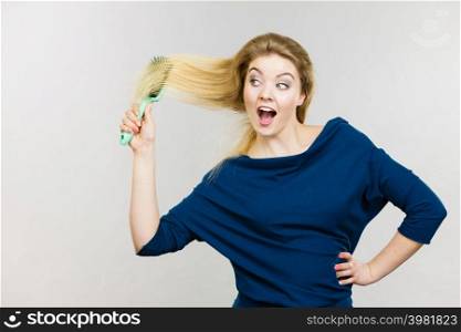Happy woman combing her hair with brush. Young smiling female with natural blond straight long hairs, studio shot on grey. Woman brushing her long hair with brush