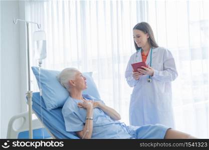 Happy woman doctor talking and using tablet to sick old female senior elderly patient lying in bed in hospital ward room in medical, technology and healthcare treatment concept. Caucasian people.